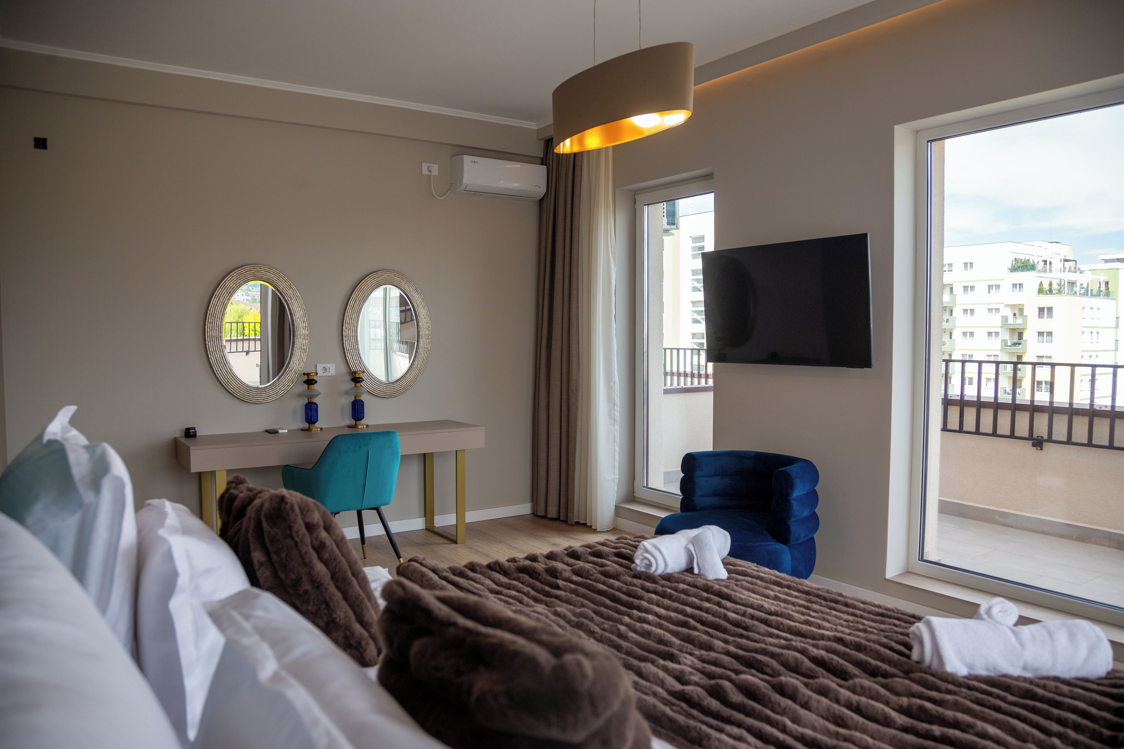 A hotel room with a bed, a desk with a chair, a TV mounted on the wall, and a window with a view of buildings.
