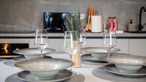 Una mesa de comedor dispuesta con platos, tazones y copas de vino, un florero con flores y electrodomésticos de cocina al fondo.