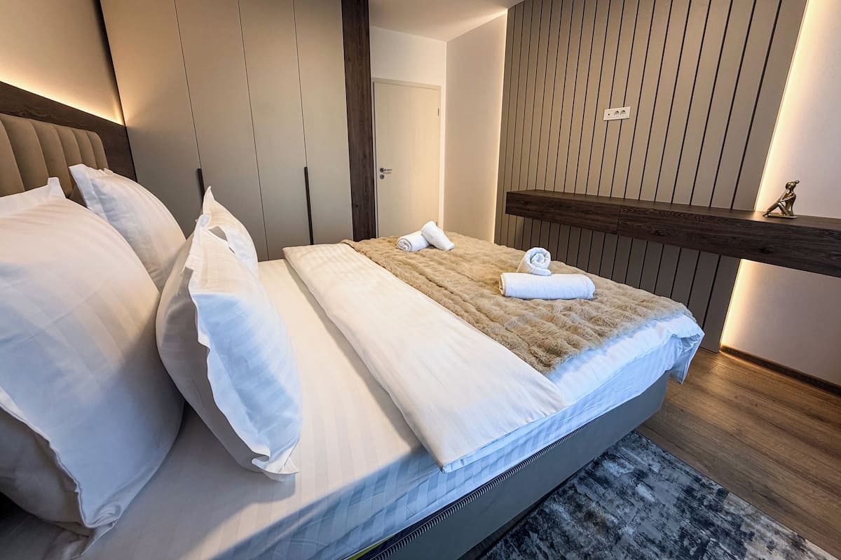 Un lit bien fait, avec du linge de lit blanc et une couverture marron, une tête de lit en bois et une étagère fixée au mur, sur laquelle repose une petite figurine.