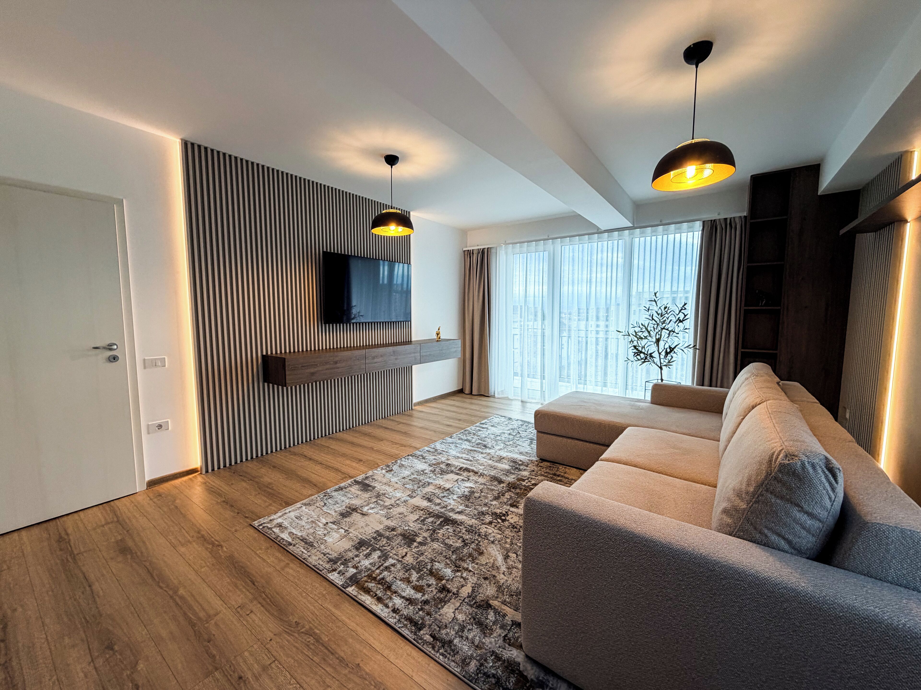 Una sala de estar moderna con un televisor de pantalla plana montado en una pared de paneles de madera, un sofá color beige, un tapete estampado y dos lámparas colgantes.