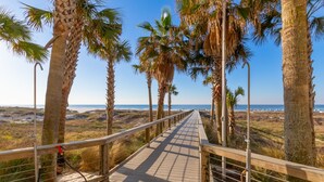 Una pasarela de madera con barandas de metal, rodeada de palmeras, que conduce a una playa con el océano al fondo.