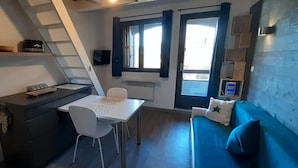 A compact room with a desk, chair, and a blue sofa. There is a window with curtains, a radiator, and a small shelf with a radio. - La Louvatière, in the heart of the village, the ideal place to discover the Monts Jura. (Mijoux)