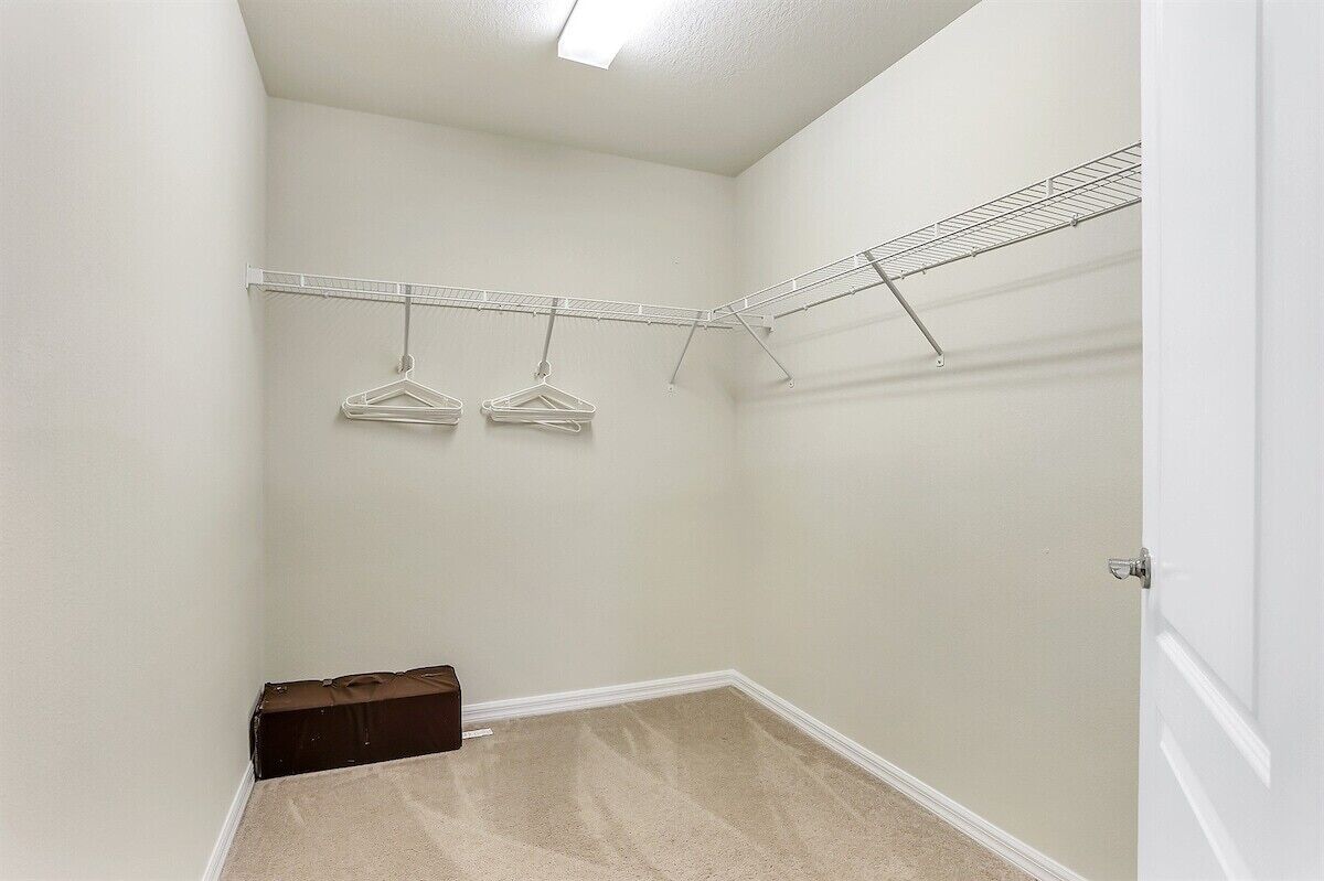 Un placard d’angle avec une étagère en fil métallique sur laquelle sont accrochés deux cintres, une valise marron posée sur le sol et une porte blanche.