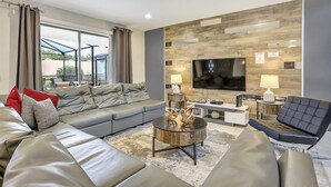 A modern living room with a grey sectional sofa, a black armchair, a round coffee table, and a flat-screen TV mounted on a wooden wall. - 2440 One of a Kind 5bd&4ba VILLA (Kissimmee)