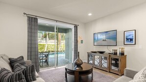 Una sala de estar moderna con un sofá, una mesa de centro y una televisión. Hay un ventanal corredizo que da a un patio con mobiliario exterior.