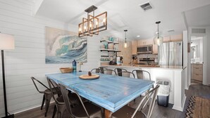 Uma sala de jantar com uma mesa azul, cadeiras e um candelabro. Cozinha visível ao fundo, com eletrodomésticos e uma geladeira.