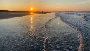 Cena de praia com um pôr do sol, águas calmas e uma pessoa caminhando pela margem.