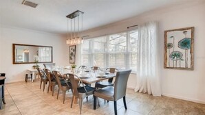 Un coin repas avec une grande table dressée pour six personnes, entourée de chaises. Un lustre est suspendu au-dessus de la table, et une fenêtre avec des stores se trouve sur le côté droit.