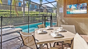 A poolside dining area with a glass enclosure, a table set for four, and a view of the pool and surrounding greenery. - 25% Off 8BR Villa! Private Pool & Spa (Kissimmee)