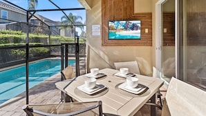 A poolside dining area with a table set for two, a TV mounted on the wall, and a view of the pool. - 25% Off 8BR Villa! Private Pool & Spa (Kissimmee)