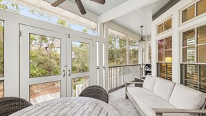 A covered porch with a wooden table and chairs, a white sofa, and large windows offering views of the surrounding greenery. - Peaceful family friendly coastal oasis with room to unwind, play, and reconnect (Watersound West)