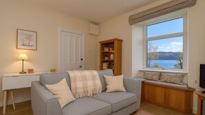 Ein Wohnzimmer mit grauem Sofa, einem Holzregal, einem Schreibtisch mit Lampe und einem Fenster mit Seeblick.