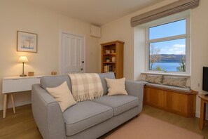 Ein Wohnzimmer mit grauem Sofa, einem Holzregal, einem Schreibtisch mit Lampe und einem Fenster mit Seeblick.