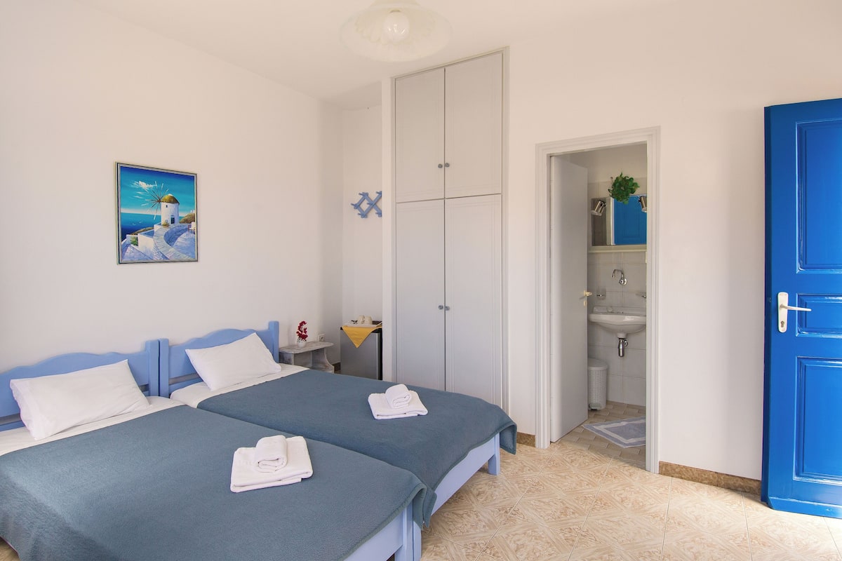 A hotel room with two beds, a painting of a coastal scene, a bathroom with a sink and mirror, and a blue door.