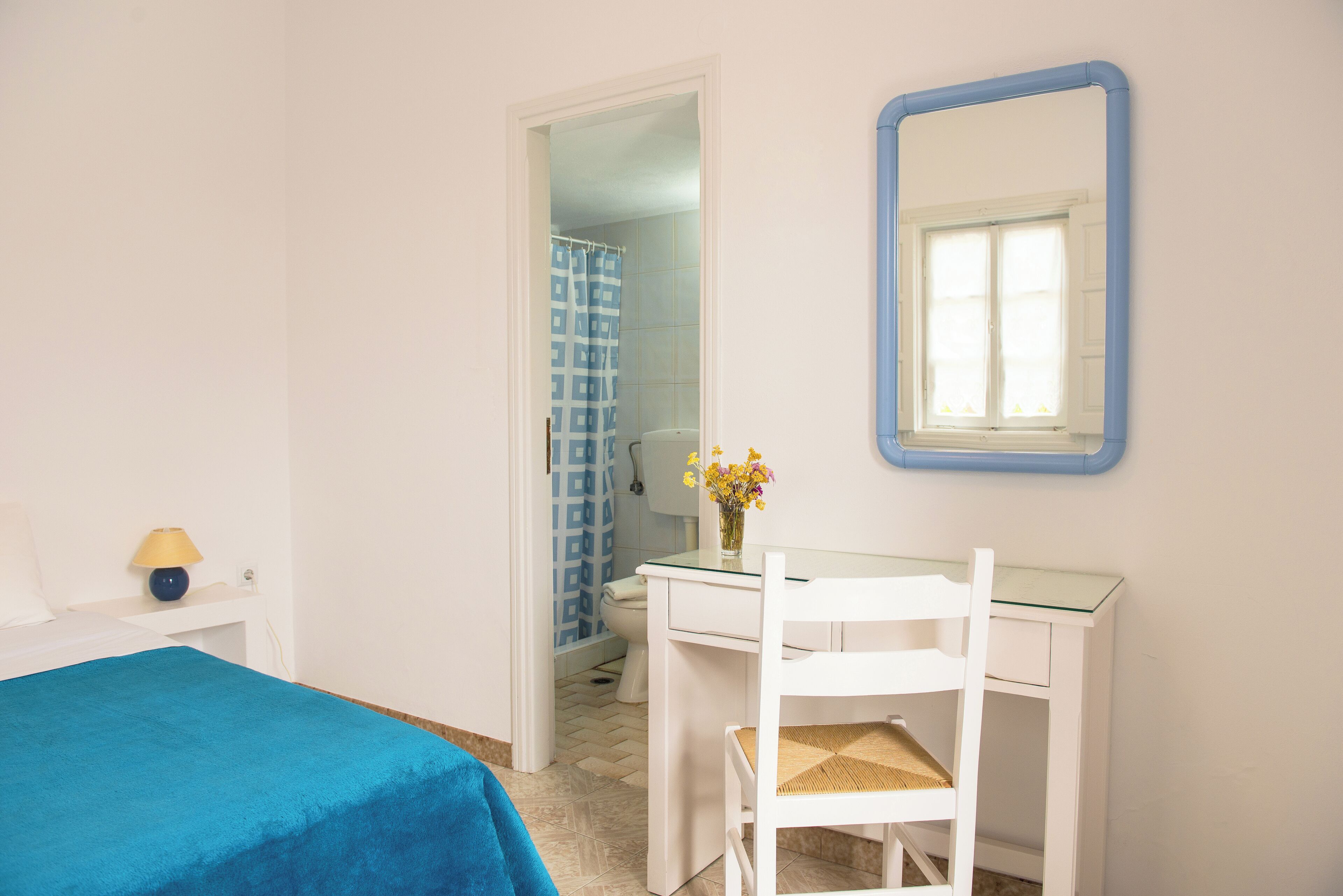 A bedroom with a bed, a desk, a chair, a mirror, and a bathroom visible through an open door.