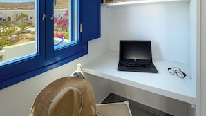 Ein Zimmer mit großem Fenster, einem Laptop auf dem Schreibtisch und einem Strohhut auf dem Boden.