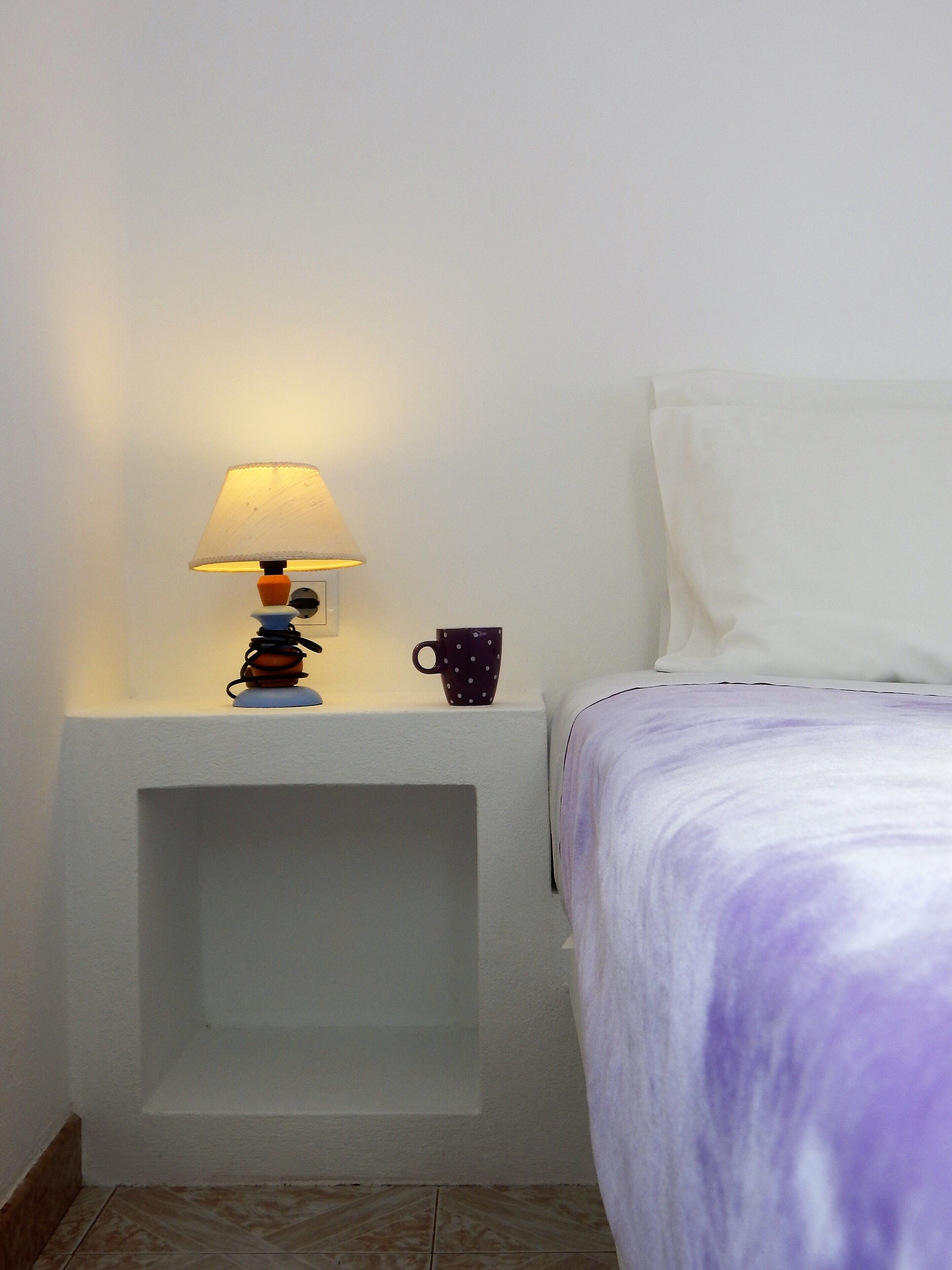 Una cama bien hecha con una colcha morada y blanca, una almohada blanca y una mesita de noche con una lámpara y una taza.