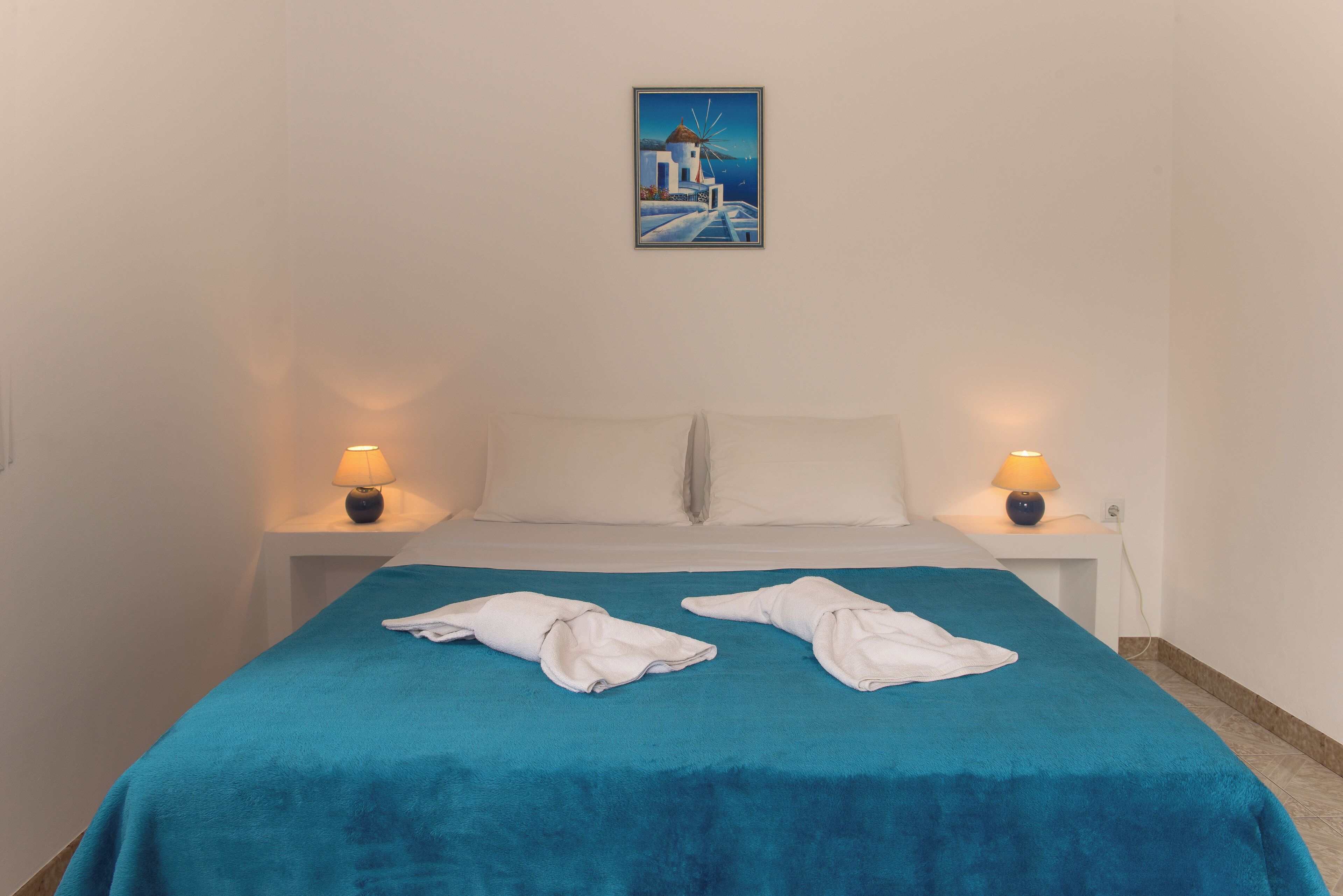 Quarto de hotel com uma cama, duas luminárias de cabeceira, um quadro na parede e duas toalhas sobre a cama.
