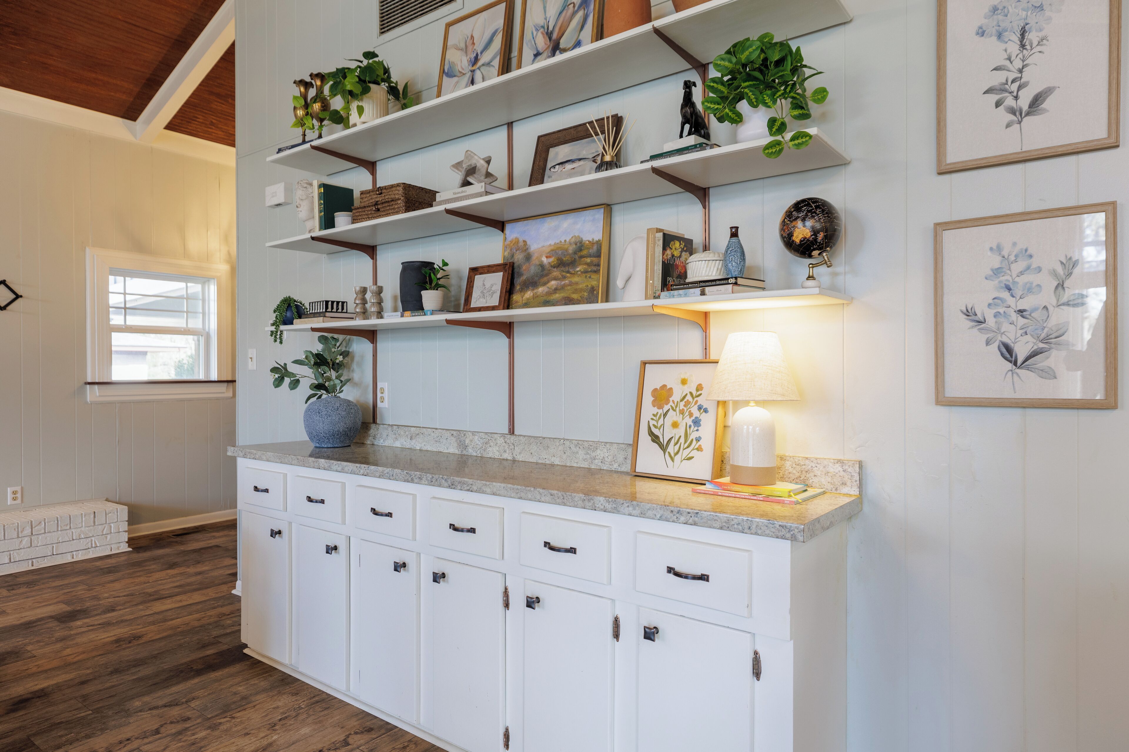 A kitchen with white cabinets, a granite countertop, and shelves with decorative items and plants.