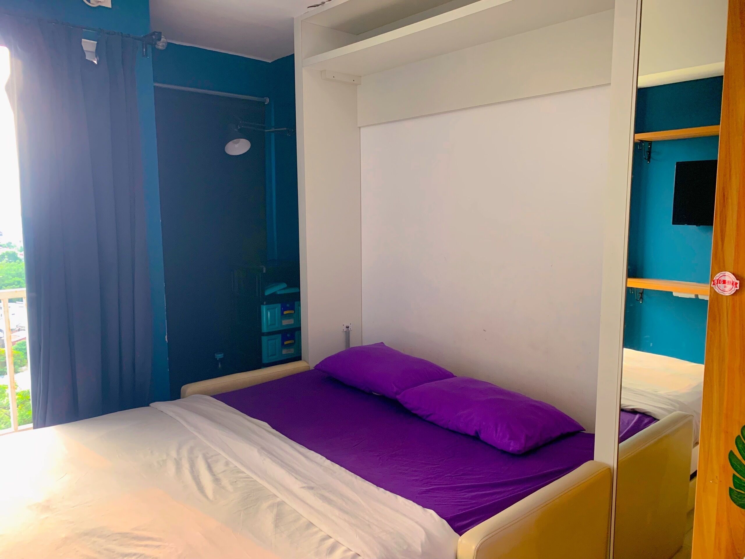 A bedroom with a bed, a window with curtains, a shower area, and a small shelf.