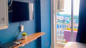 A bedroom with a TV mounted on the wall, a desk with a computer, a bed, and a balcony with a view of buildings. - Apartemen Poris 88 by Linkara (Tangerang)