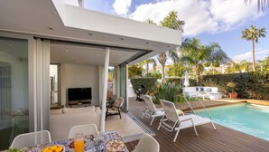 A modern outdoor living area with a pool, lounge chairs, and a dining table with a fruit basket and drinks. - Protaras Seaside Villa Narcissos 2 (Protaras)