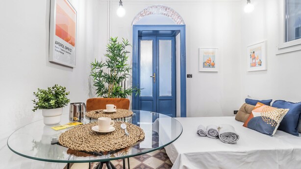 Une chambre d’hôtel compacte avec une table en verre, un lit sur lequel sont pliées des serviettes, une porte bleue et une plante en pot.