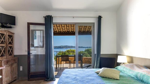 Een slaapkamer met een bed, een televisie, een houten kast, een balkon met een tafel en stoelen, en uitzicht op de zee.
