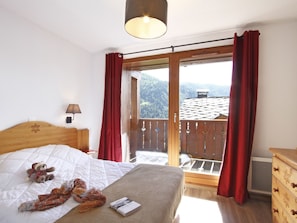 A bedroom with a bed, a teddy bear, a book, and a view of a balcony with red curtains. - Odalys Référence Le Grand Panorama 1 by Interhome (Valmeinier)