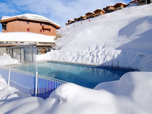 Ein Pool, umgeben von Schnee, Holzhäusern und einer Skipiste.