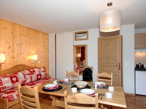 Ein gemütliches Zimmer mit Holzwand, einer Couch mit rot-weißer Musterdecke, einem Esstisch für vier Personen und einer Küchenzeile mit Kühlschrank.