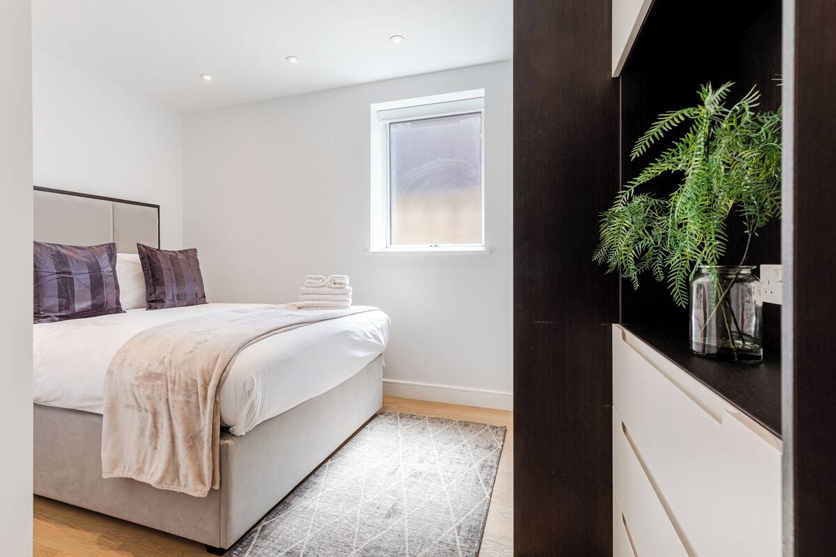 Een slaapkamer met een bed, kussens, een nachtkastje, een raam en een plant.