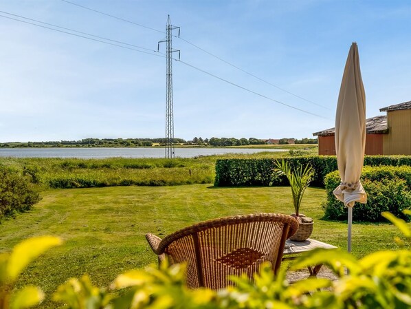 Un espace extérieur verdoyant avec une chaise en osier et un parasol, une étendue d’eau, un pylône de ligne électrique et un bâtiment en arrière-plan.