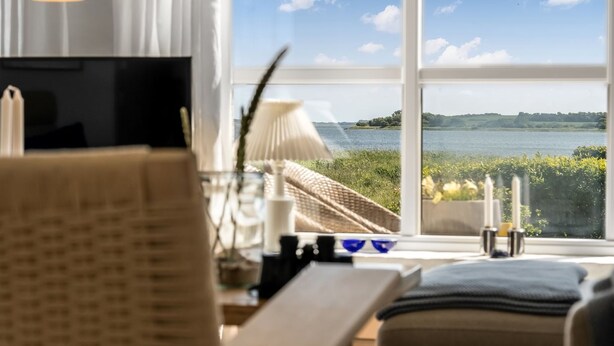 A room with a large window offering a view of a lake and distant hills. There is a chair, a table, and a lamp. - "Jurien" - 50m to the fjord by Interhome (Snedsted)