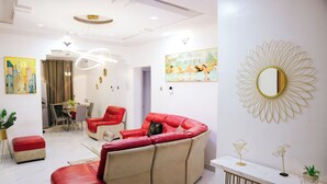 A modern living room with a red and beige sofa, a dining area with a table and chairs, and a white wall with a large mirror and artwork. - UTL Homes & Apartments
Brighton Apartment (Ibadan)