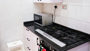 A compact kitchen with white cabinets, a microwave, a gas stove, and a small white stool. - UTL Homes & Apartments
Brighton Apartment (Ibadan)
