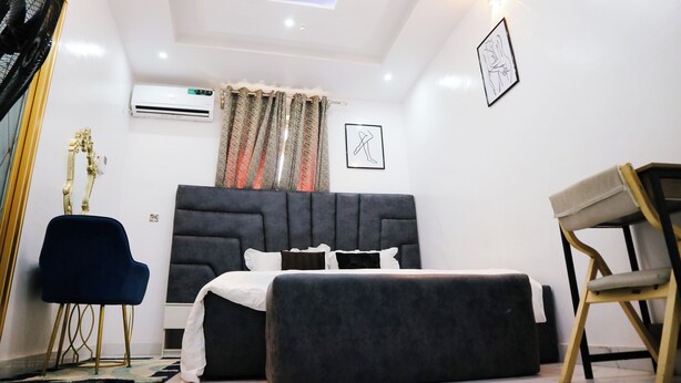 A modern hotel room with a large bed, a bedside table, a chair, an air conditioning unit, and a ceiling light fixture. - UTL Homes & Apartments
Brighton Apartment (Ibadan)
