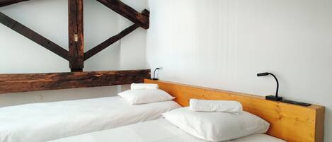Una habitación con dos camas individuales, vigas de madera, paredes blancas y una mesita de noche sencilla con lámpara.