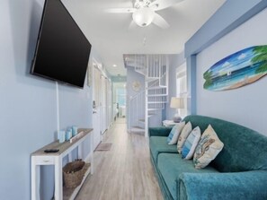 Ein Zimmer mit an der Wand montiertem Fernseher, einem grünen Sofa mit Dekokissen, einem weißen Konsolentisch und einem Deckenventilator.
