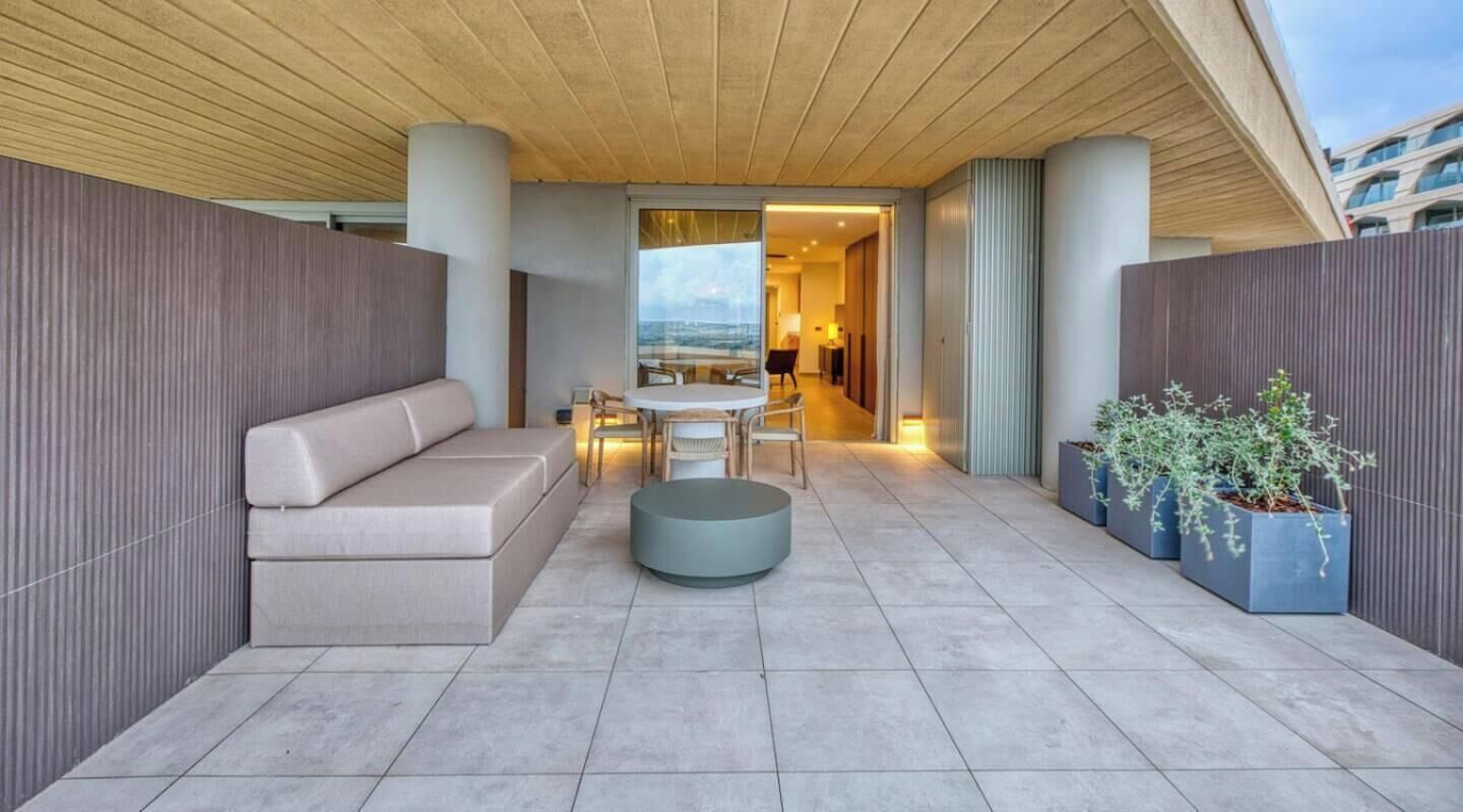 Un espace extérieur moderne aménagé avec un canapé, une table ronde et des chaises, entouré de plantes en pot et offrant une vue sur l’océan.