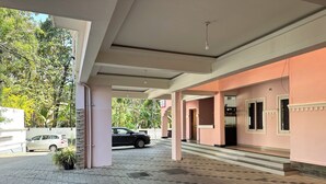 A covered outdoor area with a tiled floor, a car parked nearby, and a building with pink walls and a door. - 2BHK Apartment near Guruvayur Temple (Guruvayur)
