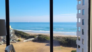 Une vue d’une plage avec des vagues de l’océan, un ciel dégagé et un bâtiment doté de nombreuses fenêtres.
