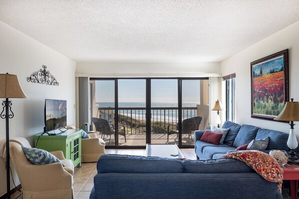 Uma sala de estar com sofá, cadeiras, televisão, um armário verde e uma varanda com vista para o oceano.