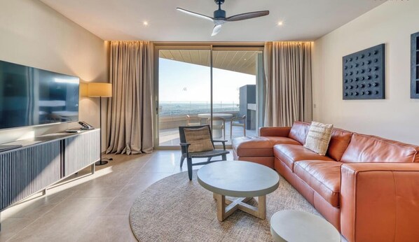 Uma sala de estar moderna com sofá de couro, mesa de centro redonda, TV de tela plana e uma grande janela com vista para o mar.