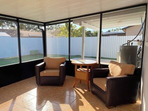 Une terrasse ensoleillée avec deux fauteuils confortables et une petite table, de grandes fenêtres et un réservoir d’eau en arrière-plan.