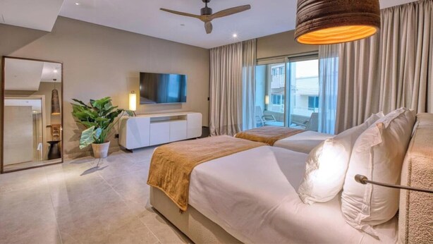 Une chambre d’hôtel moderne dotée d’un grand lit, d’une télévision, d’un ventilateur de plafond et d’un balcon avec des rideaux.