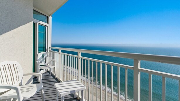 Un balcon avec des chaises blanches et une table, donnant sur une plage et l’océan.