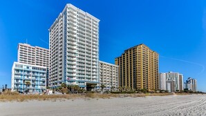 Un hôtel situé en bord de mer, composé de plusieurs immeubles de grande hauteur, avec une plage de sable fin et un ciel d’un bleu éclatant.