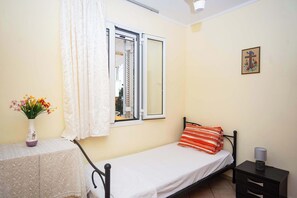 A small bedroom with a single bed, a bedside table, a nightstand, a window with curtains, and a wall-mounted picture. - Spacious 2 Bd Detached House in Kefalonia w Patio (Lixouri)