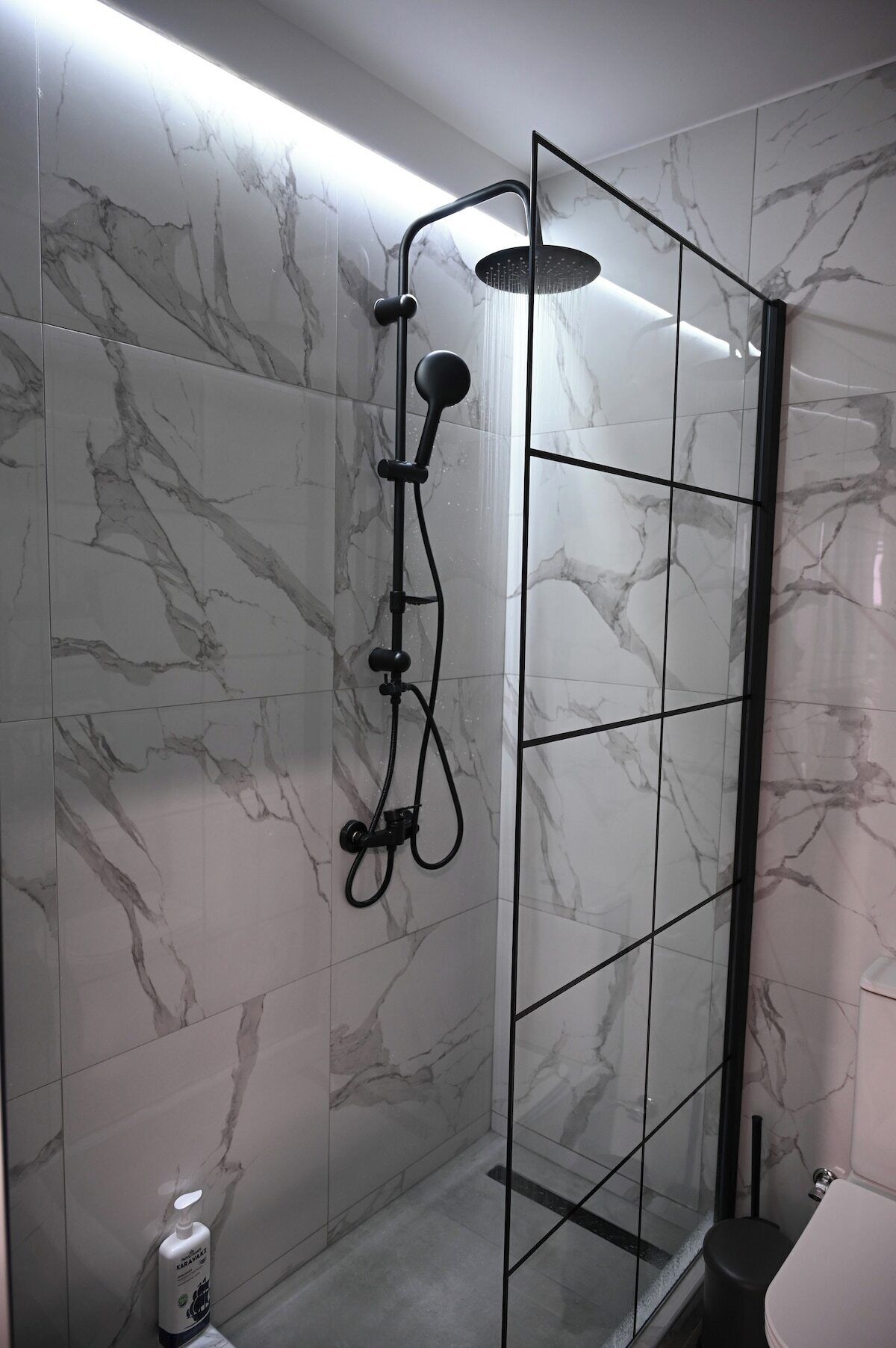 Een moderne douchecabine met een glazen deur, wanden met een marmerlook en een handdouche.